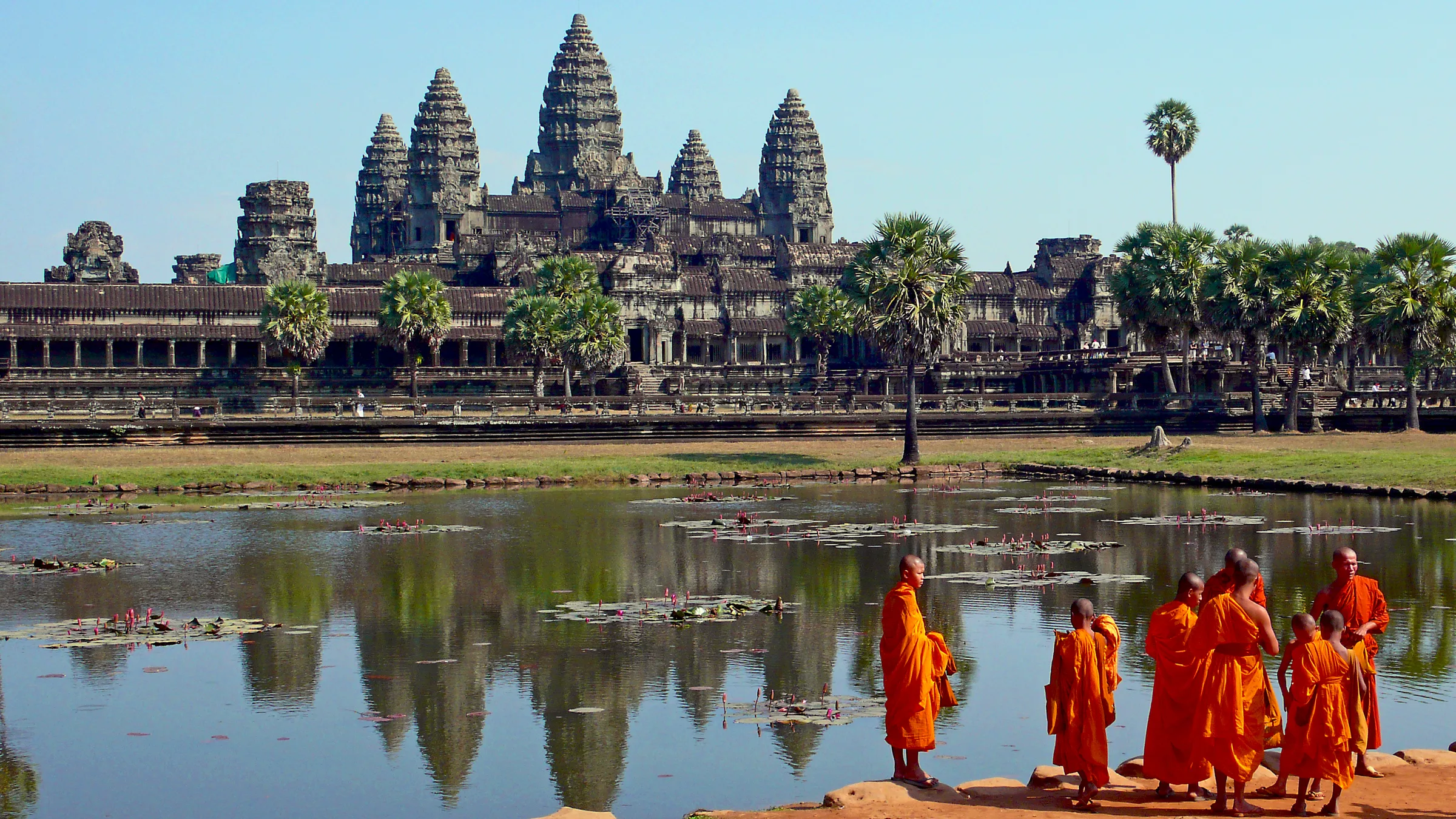 Angkor Wat'ın su birikintisine yansıyan beş kuleli silüeti; önünde turuncu cübbeli Budist keşişler yürüyor