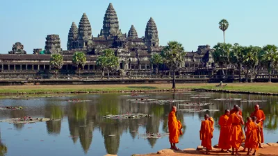 The five-towered silhouette of Angkor Wat reflected in a pool of water, with orange-robed Buddhist monks walking in front