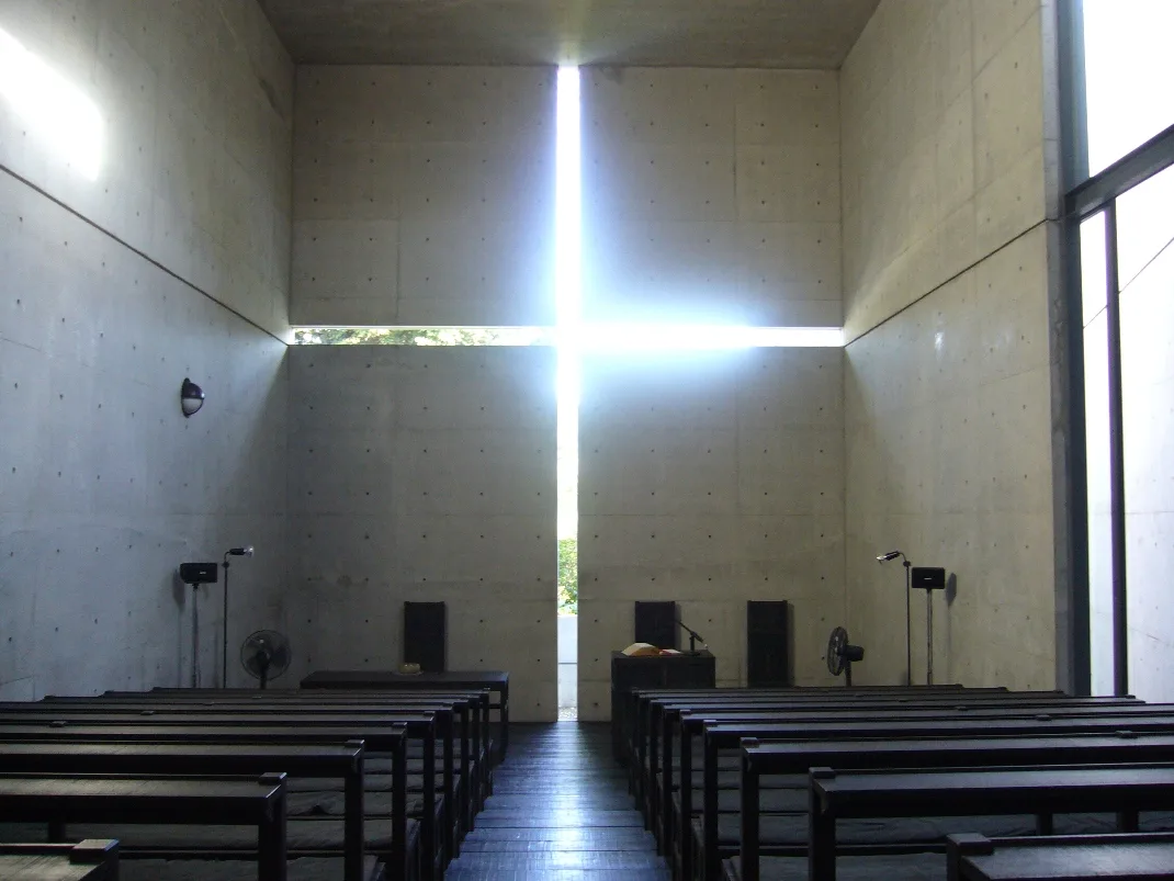 In the dim exposed-concrete interior of the Church of the Light, a bright cross of light entering through the cross-shaped slit in the wall behind the altar