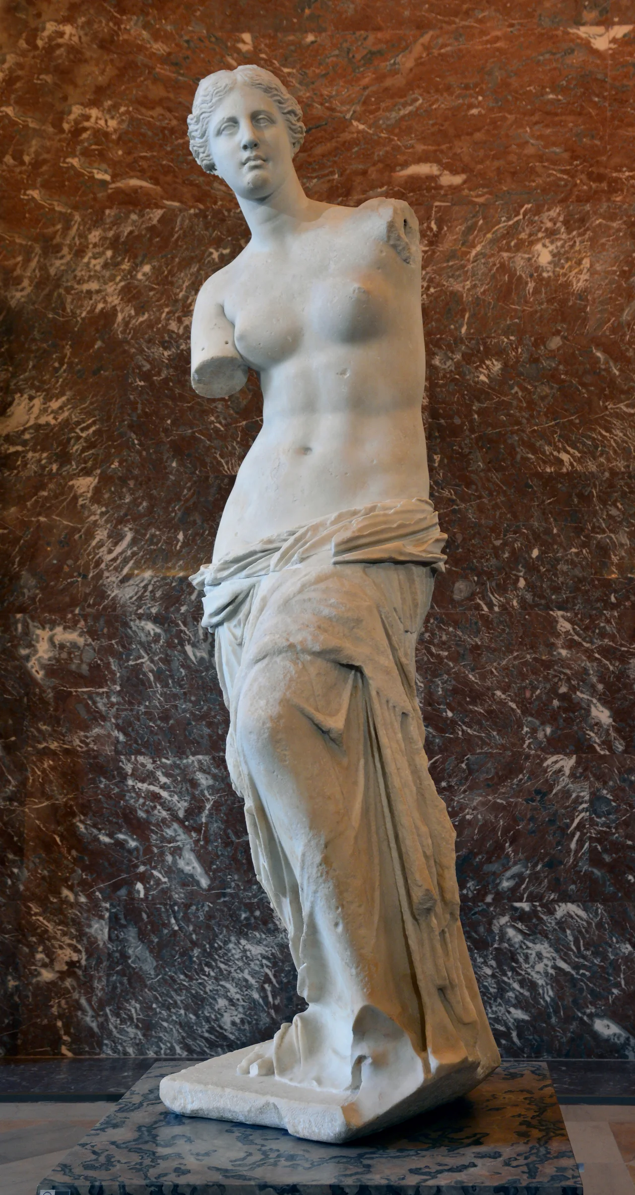 Front view of the Venus de Milo: a female figure with missing arms, bare upper body, lower body wrapped in marble drapery