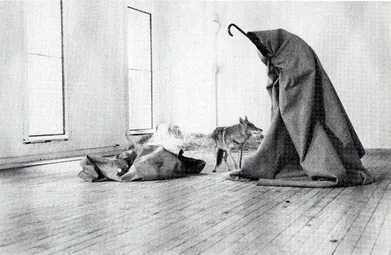 Joseph Beuys wrapped in a felt blanket holding a staff in a gallery room, with a wild coyote standing nearby looking at him