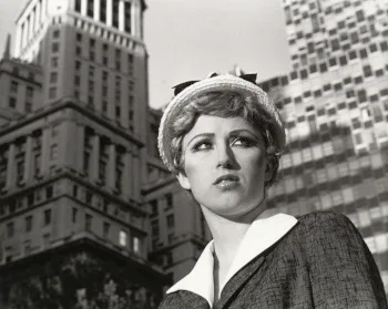 Untitled Film Still #21: a black-and-white close view of a young woman in a suit and hat looking upward in front of city buildings