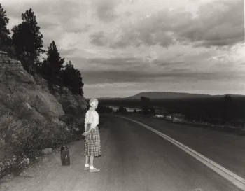 Untitled Film Still #48: a small figure with a suitcase at the edge of an empty country road, looking into the distance