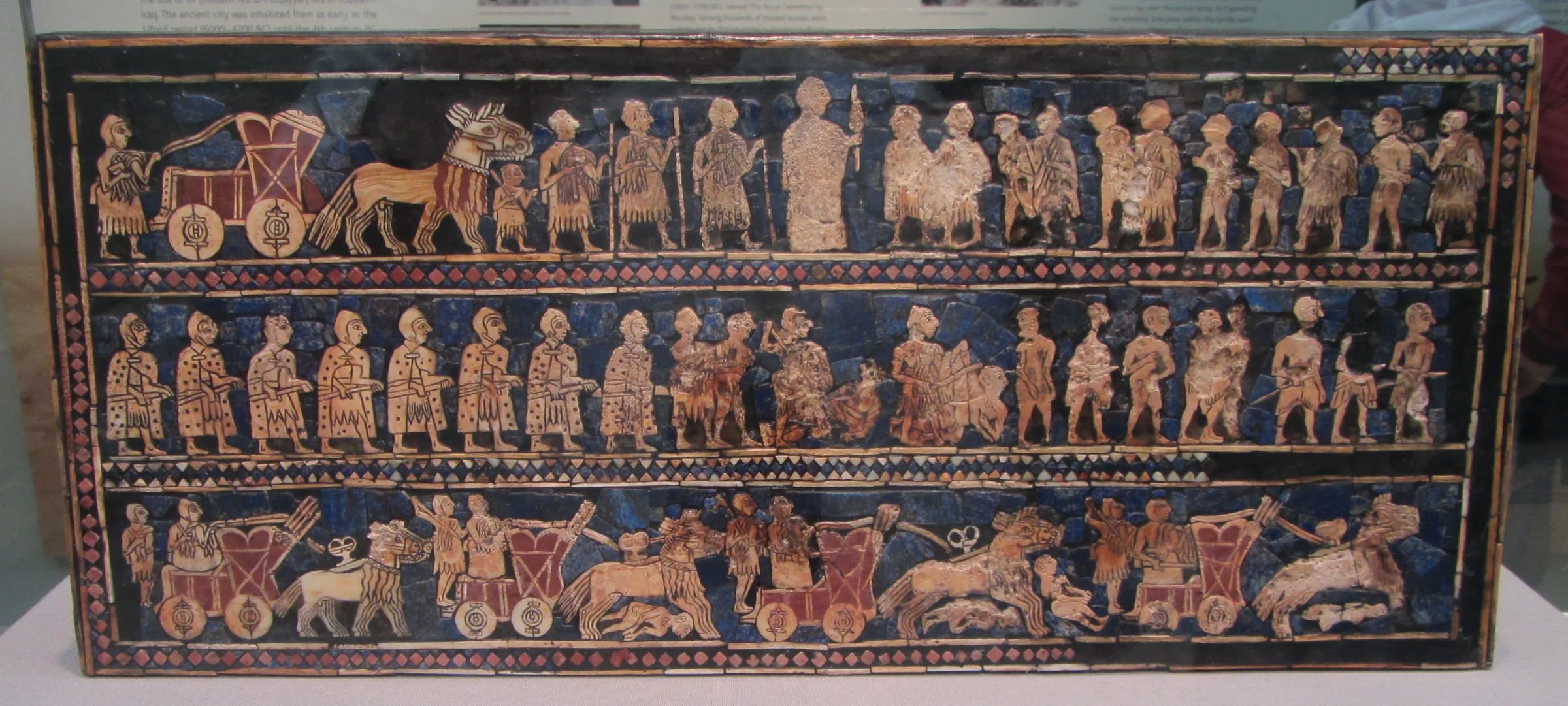 Rectangular Sumerian panel inlaid in blue and shell, in registers; wheeled wagons appear in the lower rows