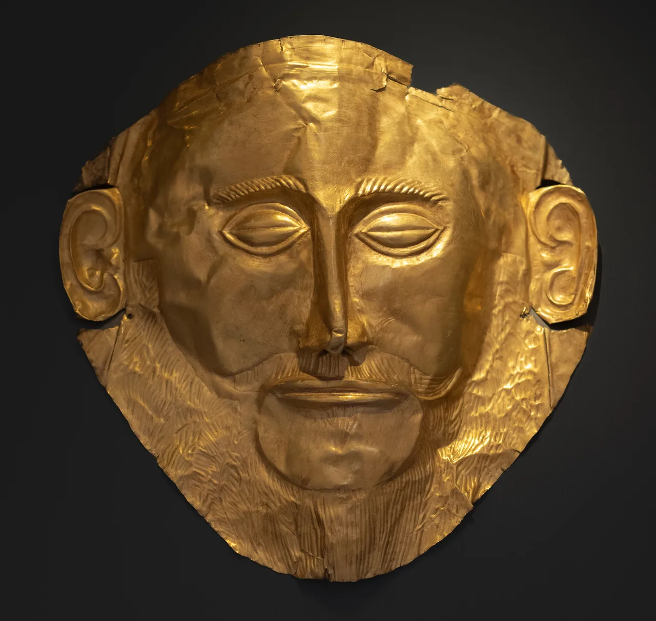 The gold death mask displayed at the National Archaeological Museum in Athens — a stylized bearded face hammered from thin gold sheet