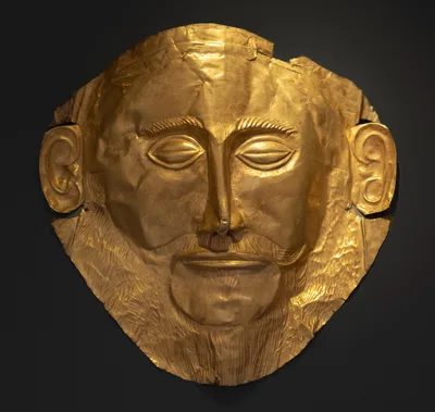 The gold death mask displayed at the National Archaeological Museum in Athens — a stylized bearded face hammered from thin gold sheet