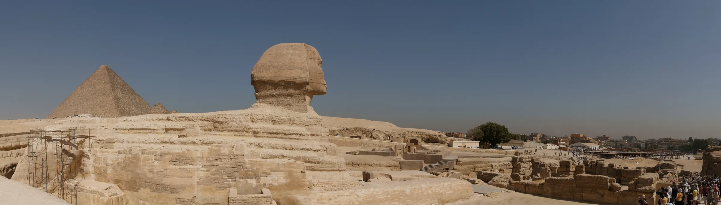 Önde Büyük Sfenks'in geniş heykeli, arkasında çöl üzerinde yükselen Giza piramitlerinin panoramik görüntüsü