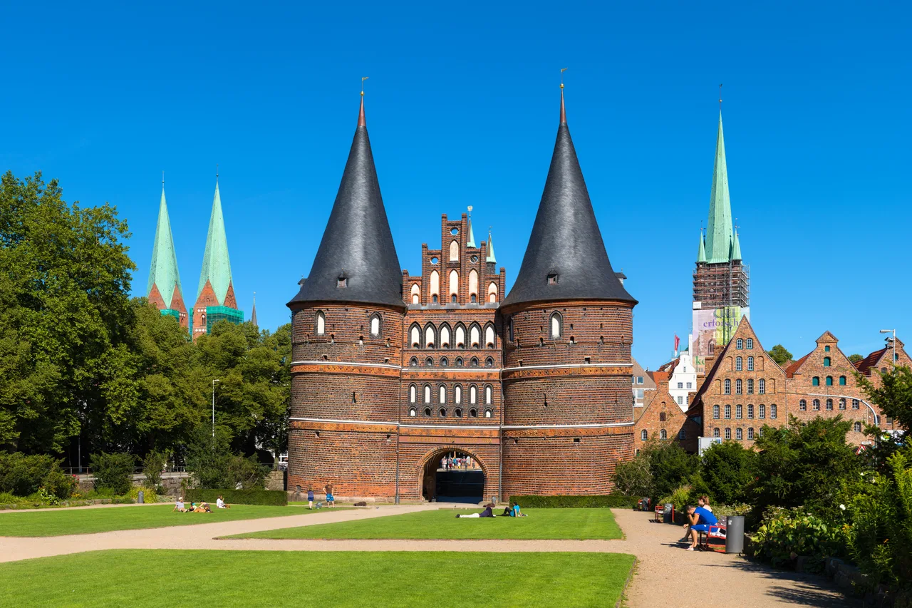 Lübeck's Holstentor gate: a late-Gothic red-brick city gate with two round towers capped with pointed tiled spires, set behind a green square under a blue sky