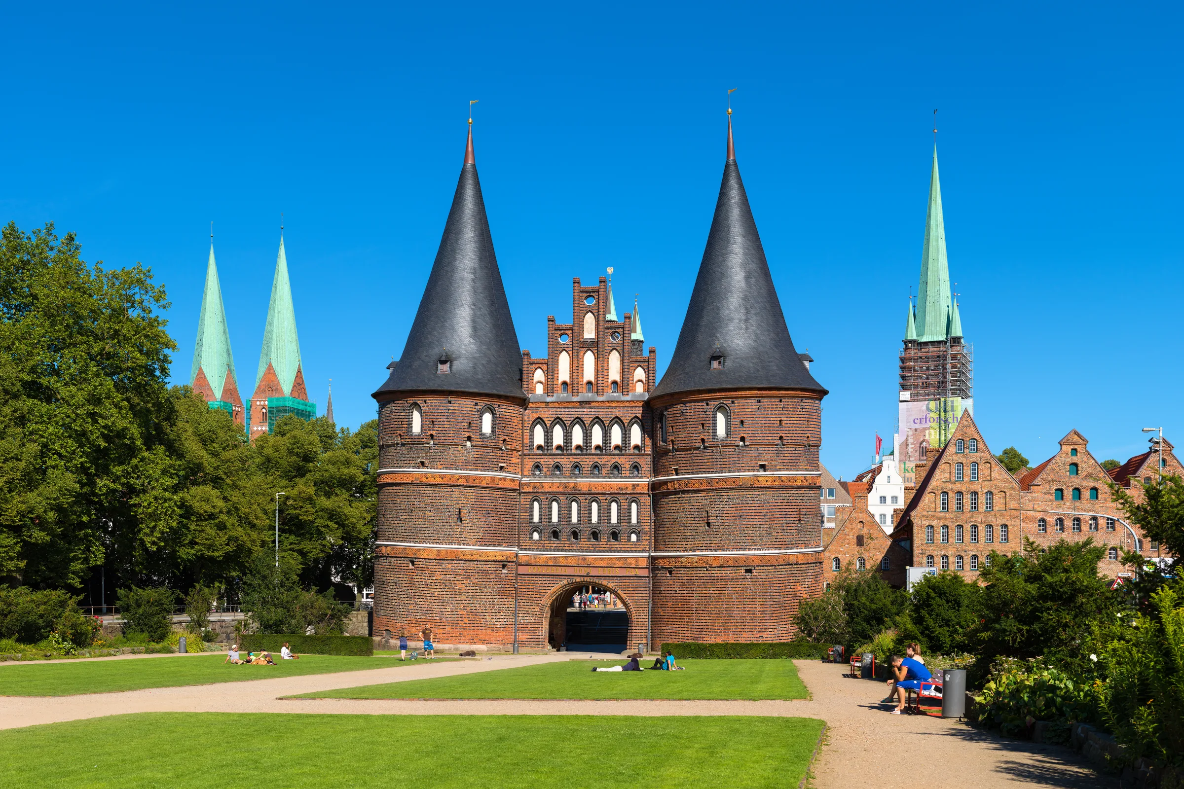 Lübeck'in Holstentor kapısı: iki yuvarlak silindirik kuleli, kırmızı tuğlalı, sivri kiremit külahlı geç-gotik şehir kapısı, önündeki yeşil meydan ve mavi gökyüzüyle
