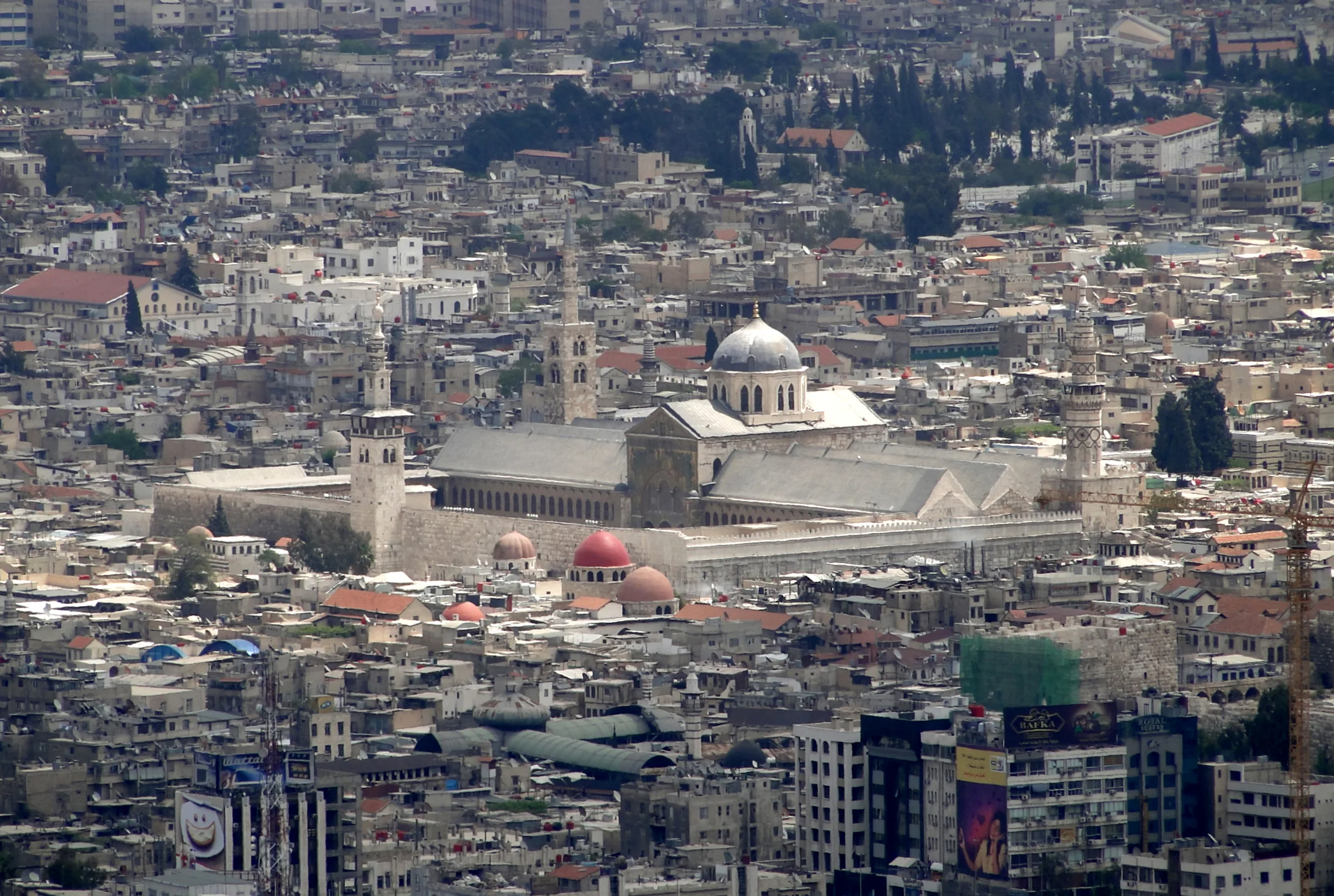 Şam'daki Emevi Camii'nin geniş iç avlusu; karşıda altın ve renkli taş mozaiklerle bezeli ana ibadet binasının üç kemerli cephesi ve uzun minareleri, ön planda mermer döşeli avlu