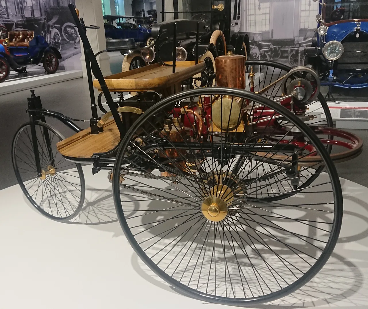 Modern replica of the Benz Patent-Motorwagen: a three-wheeled open vehicle with wood-and-metal frame, a horizontal single-cylinder engine, and a large horizontal flywheel at the rear axle