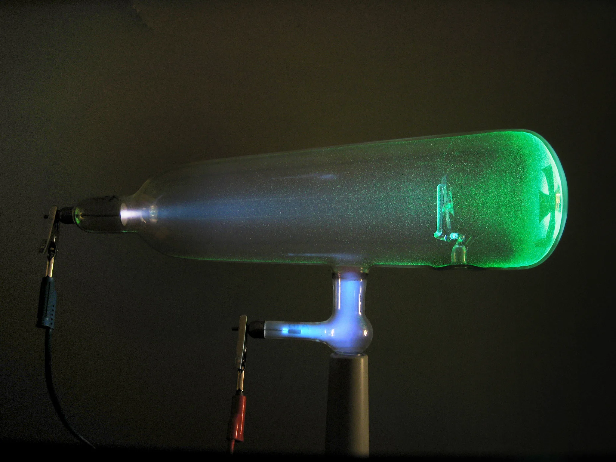 A Crookes tube in operation: a greenish glow inside the glass and the shadow of a cross-shaped obstacle