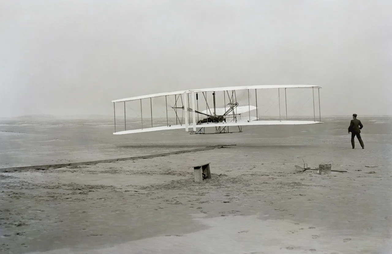 Black-and-white photograph from 17 December 1903: the Wright Flyer airborne a few metres above the ground, pilot lying flat on the lower wing; Wilbur visible at right running by the wingtip