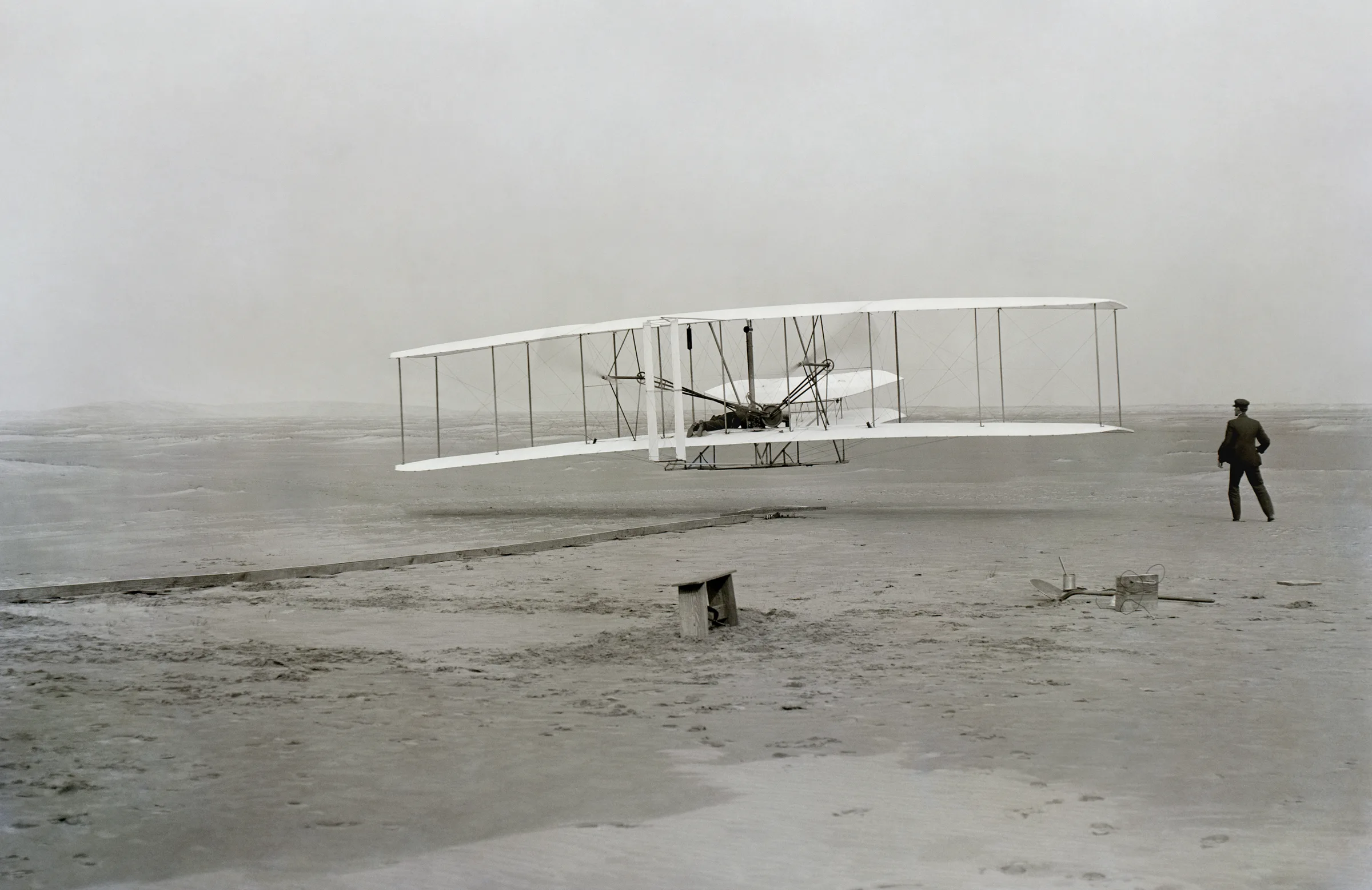 17 Aralık 1903'te çekilmiş siyah-beyaz fotoğraf: Wright Flyer havada birkaç metre yükseklikte, pilot yatay olarak alt kanada uzanmış; sağda Wilbur kanat ucunda koşarken görülüyor