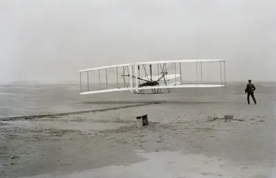 Black-and-white photograph from 17 December 1903: the Wright Flyer airborne a few metres above the ground, pilot lying flat on the lower wing; Wilbur visible at right running by the wingtip