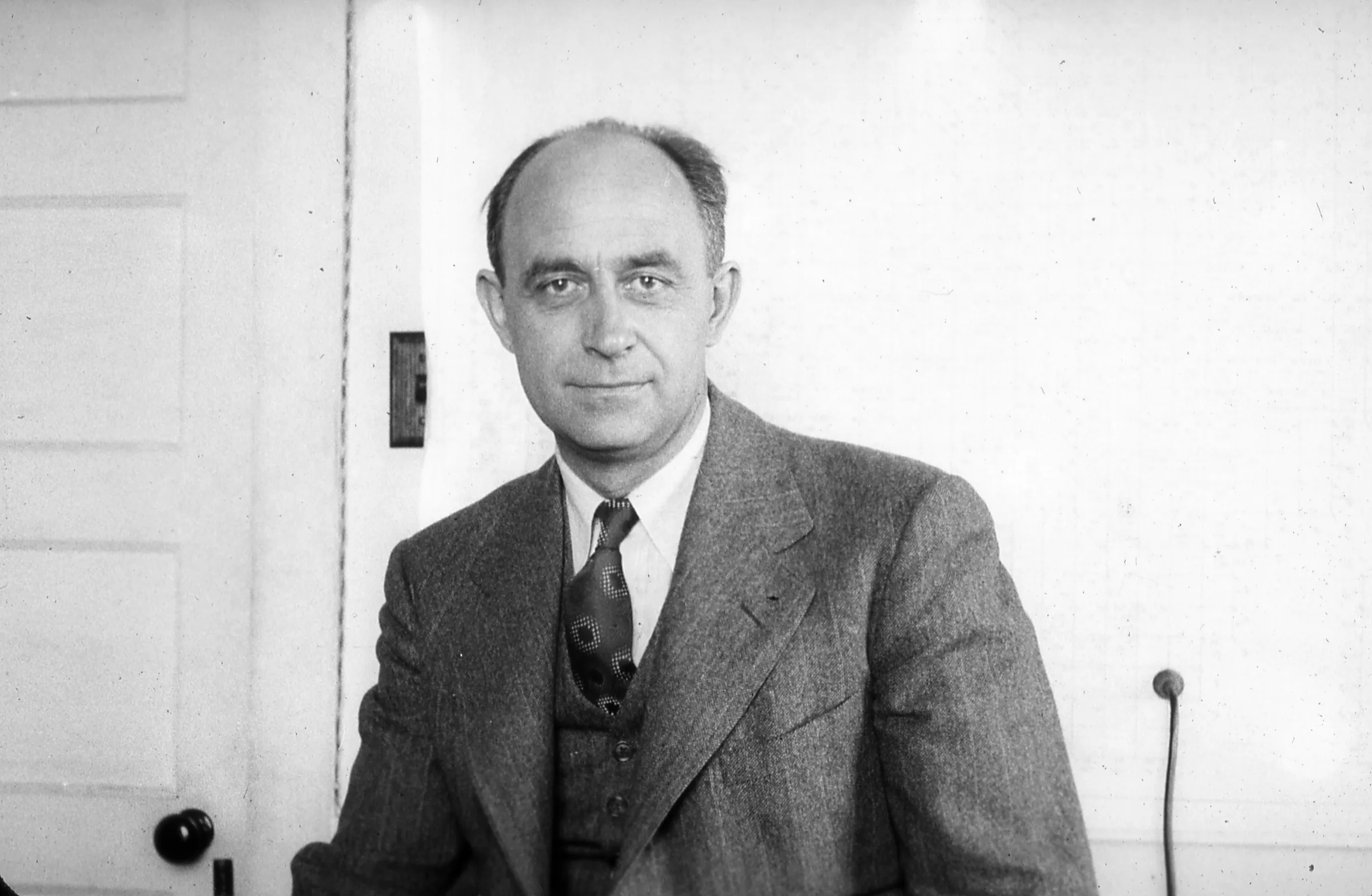 An informal black-and-white portrait of Enrico Fermi: a middle-aged Italian physicist in a white shirt, smiling, looking obliquely at the camera