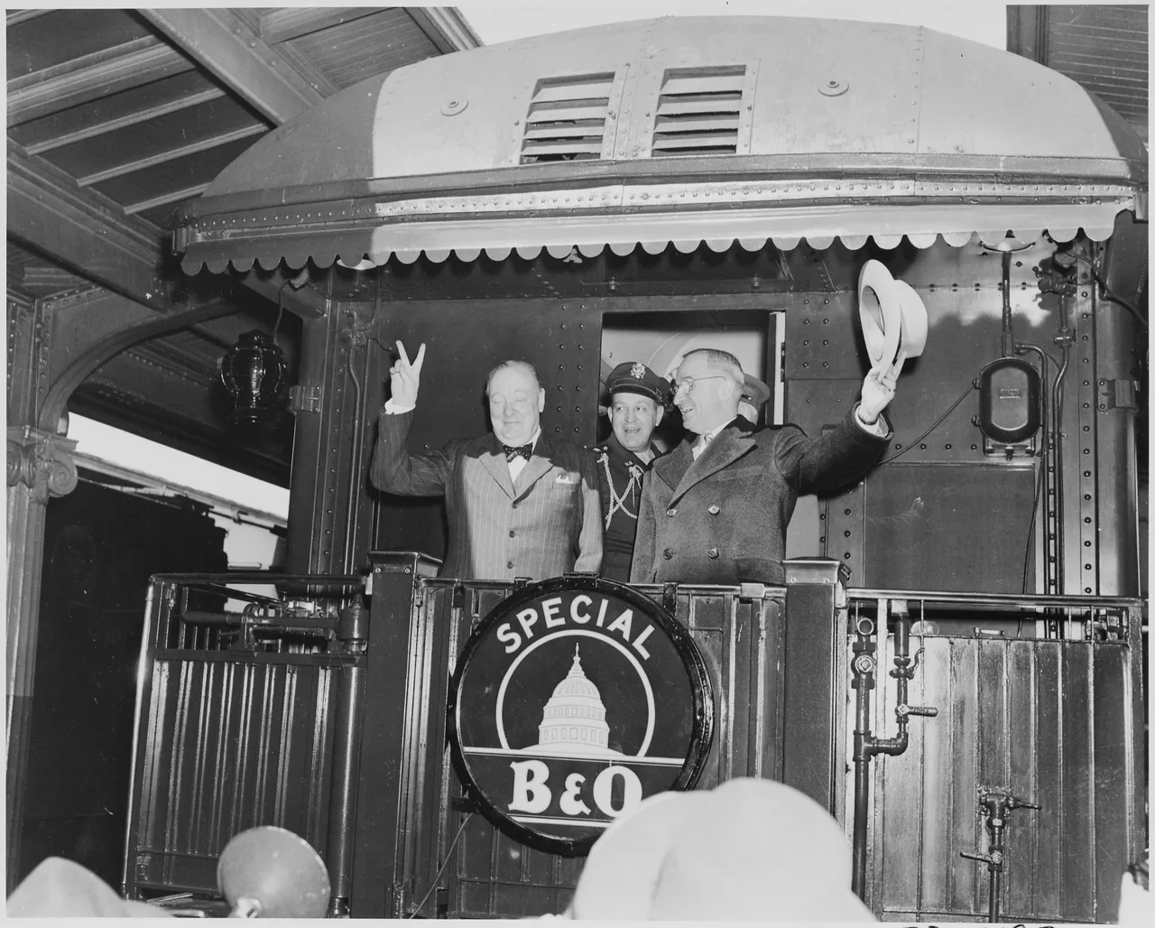 Black-and-white March 1946 photograph: on the rear platform of a train, President Truman waves his hat while Winston Churchill flashes the two-finger V-for-Victory sign; General Vaughan stands behind