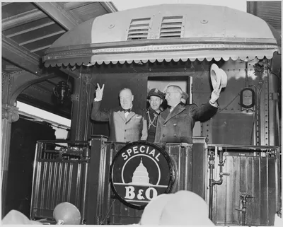 Black-and-white March 1946 photograph: on the rear platform of a train, President Truman waves his hat while Winston Churchill flashes the two-finger V-for-Victory sign; General Vaughan stands behind