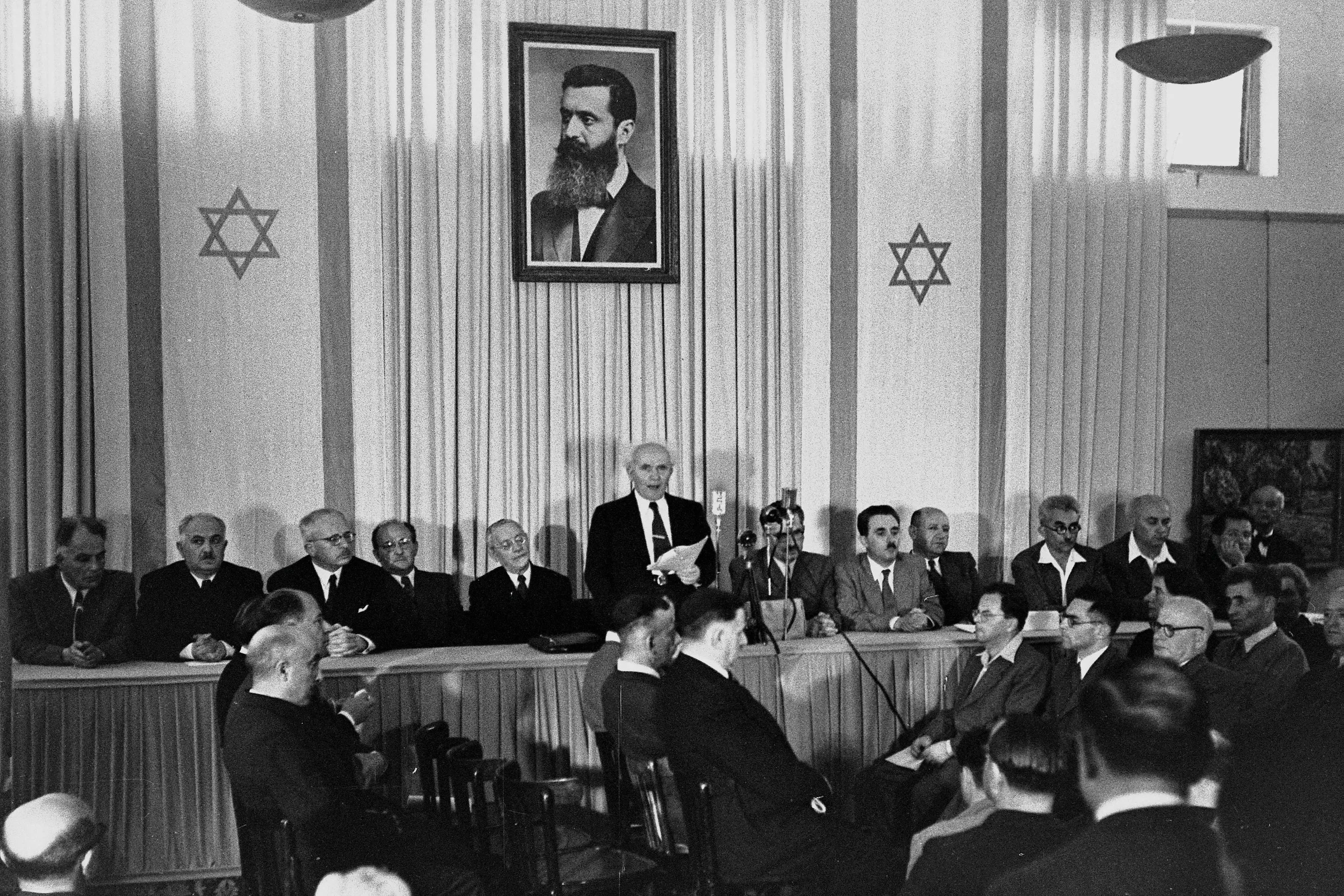 14 Mayıs 1948 Tel Aviv: David Ben-Gurion uzun bir masanın arkasında ayakta, elindeki belgeyi okuyor; arkasındaki duvarda Theodor Herzl'in büyük portresi ve iki İsrail bayrağı asılı; masanın iki yanında oturan üyeler ona bakıyor