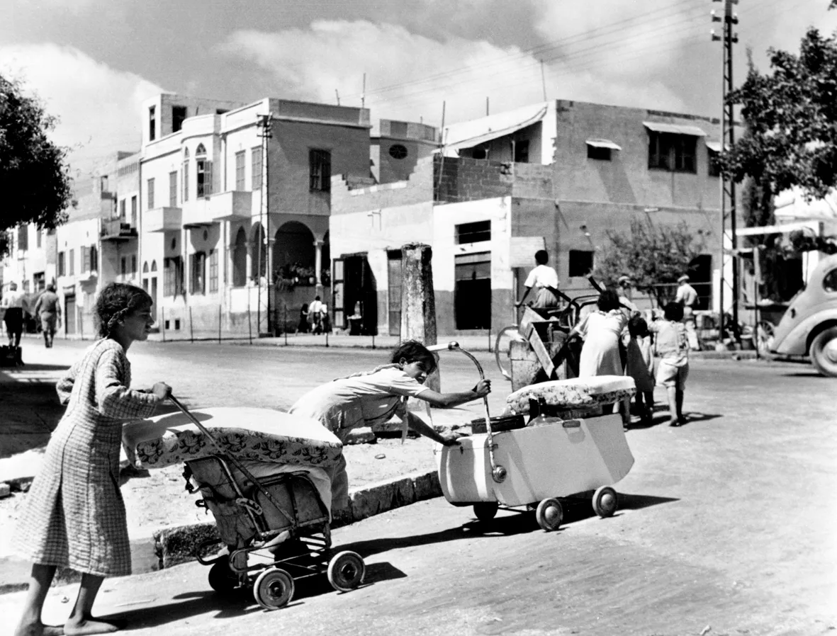 1948: Yafa'dan ayrılan iki Filistinli genç kız bebek arabaları ve el arabalarına yükledikleri eşyalarını itiyor; arka planda boş cadde