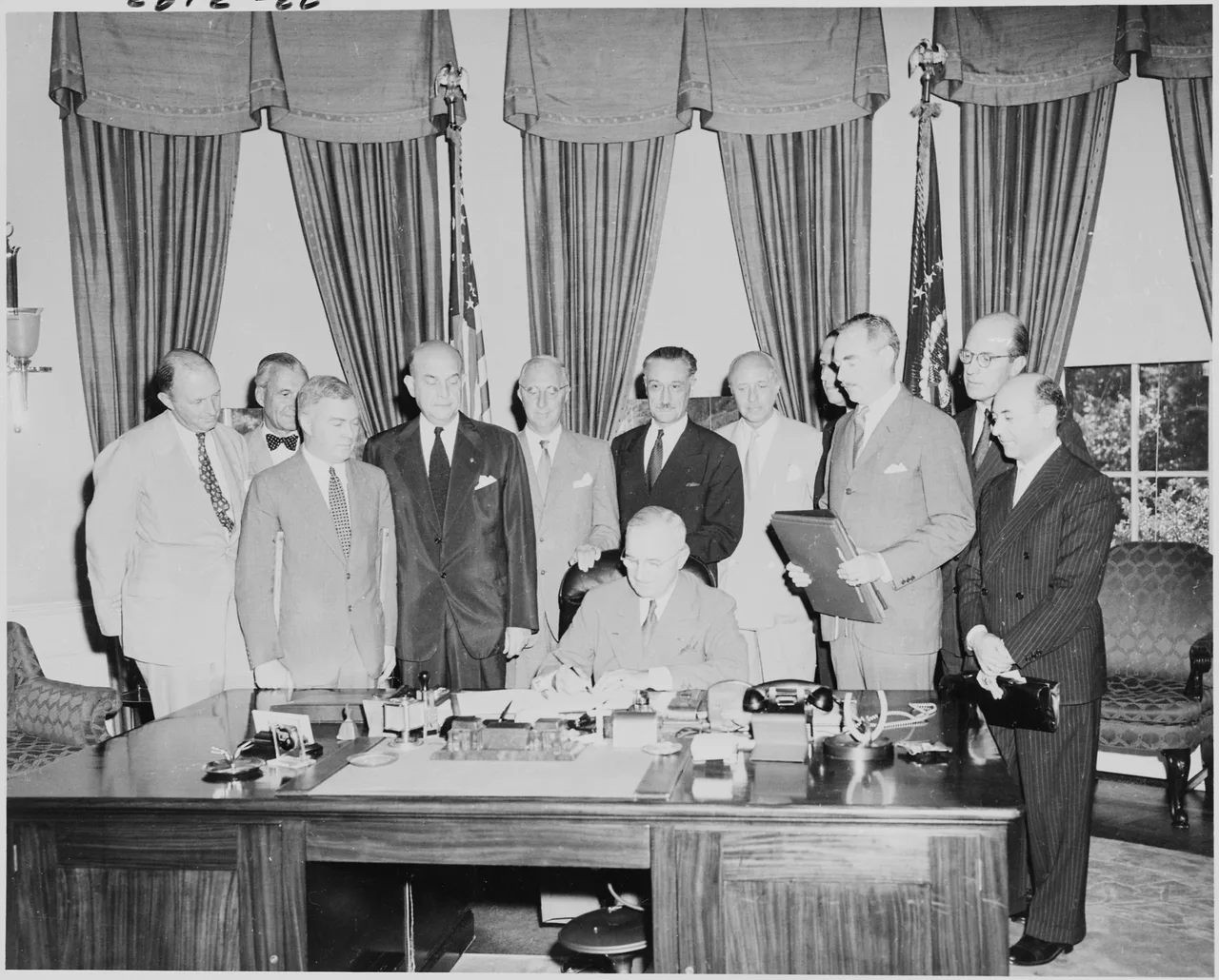 In August 1949 in the Oval Office, President Truman is seated at his desk signing a document; around him a row of European and American diplomats in suits stands looking on