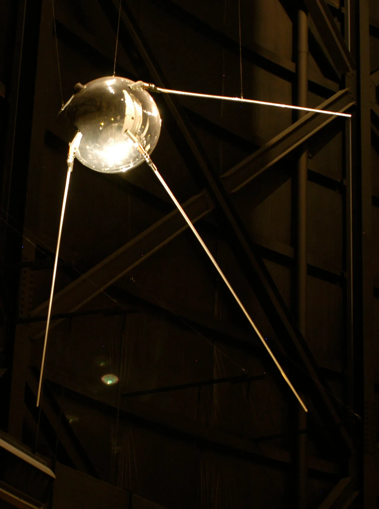 Bir müze vitrininde sergilenen Sputnik 1 modeli; siyah arka planda parlak küre ve geriye doğru uzanan dört anten net biçimde aydınlatılmış