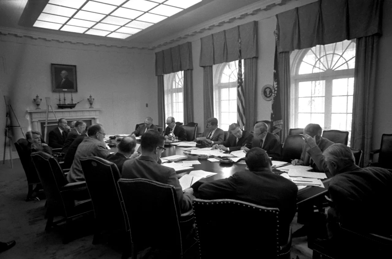 Black-and-white 1962 photograph: men in dark suits seated around a long cabinet table, some turned in discussion, others looking at documents; Kennedy at one side of the table