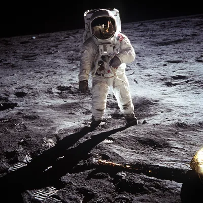 Buzz Aldrin in his white Apollo space suit stands alone on the grey lunar surface; the visor of his helmet reflects the lunar module and Armstrong, who is taking the photograph