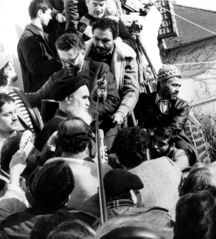17 Ocak 1979 tarihli siyah-beyaz fotoğraf: Paris yakınındaki Neauphle-le-Château'da Humeyni siyah cübbesi ve siyah sarığıyla küçük bir bahçede oturmuş gazetecilere konuşuyor; etrafında mikrofonlar ve kameralar