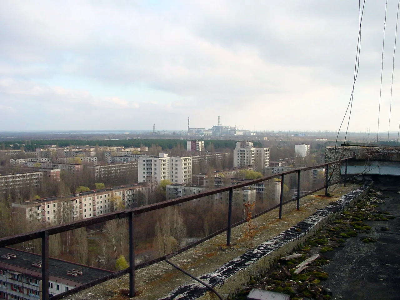 2005'te boş bir Pripyat apartmanının çatısından çekilmiş geniş açılı renkli fotoğraf: önde terk edilmiş binaların düz çatıları, arkada ağaçlar ve uzakta Çernobil santralinin baca ve reaktör binası siluet hâlinde