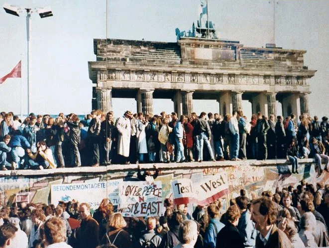 East and West Germans gathered on top of the wall at the Brandenburg Gate in 1989 — a crowd waving a flag, shaking hands, smiling
