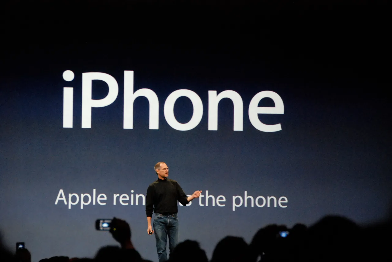 Steve Jobs on stage in his black T-shirt and blue jeans, holding up the first iPhone as he introduces it — a large projection of the device behind him