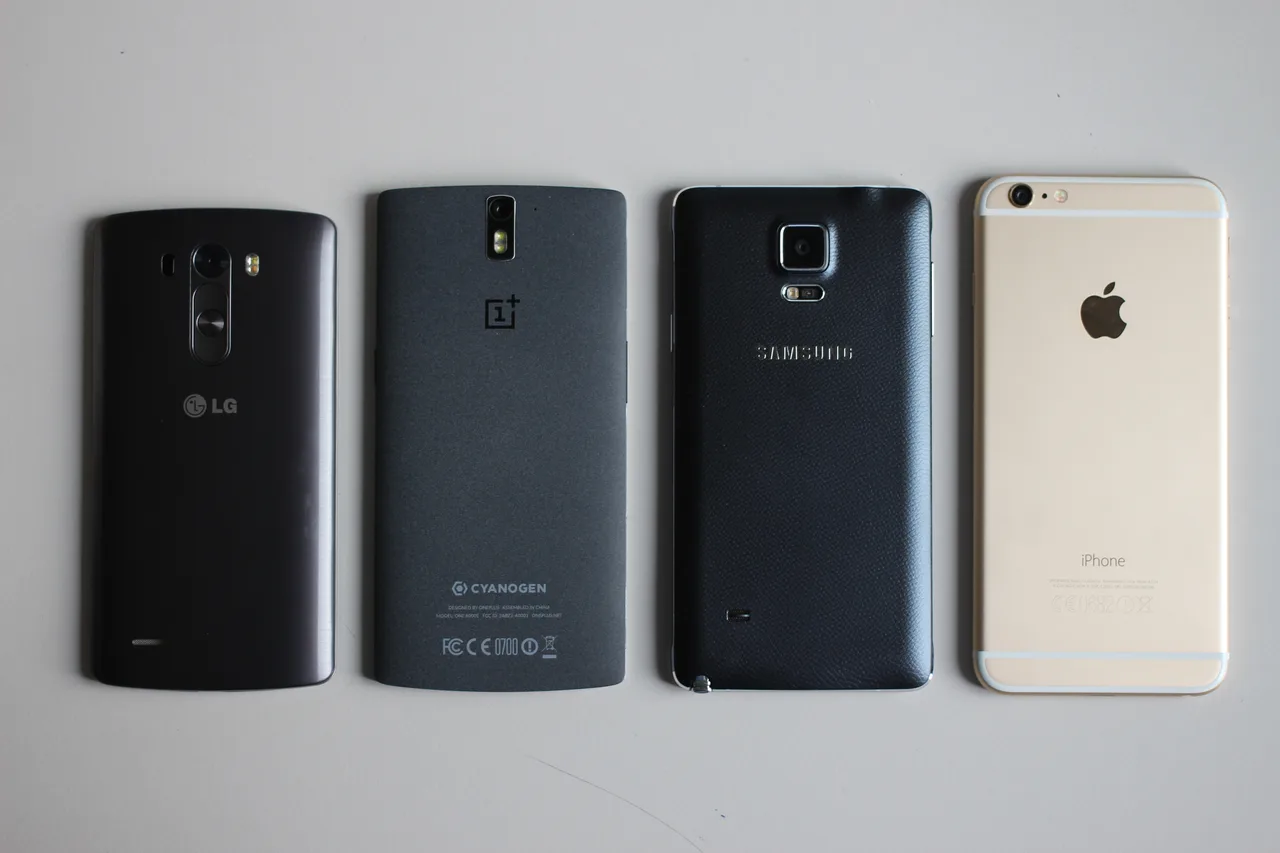 Four smartphones back-side-up on a white surface: OnePlus One, LG G3, iPhone 6 Plus and Samsung Galaxy Note 4 — each thin, camera-equipped, glass-and-metal.