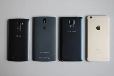 Four smartphones back-side-up on a white surface: OnePlus One, LG G3, iPhone 6 Plus and Samsung Galaxy Note 4 — each thin, camera-equipped, glass-and-metal.