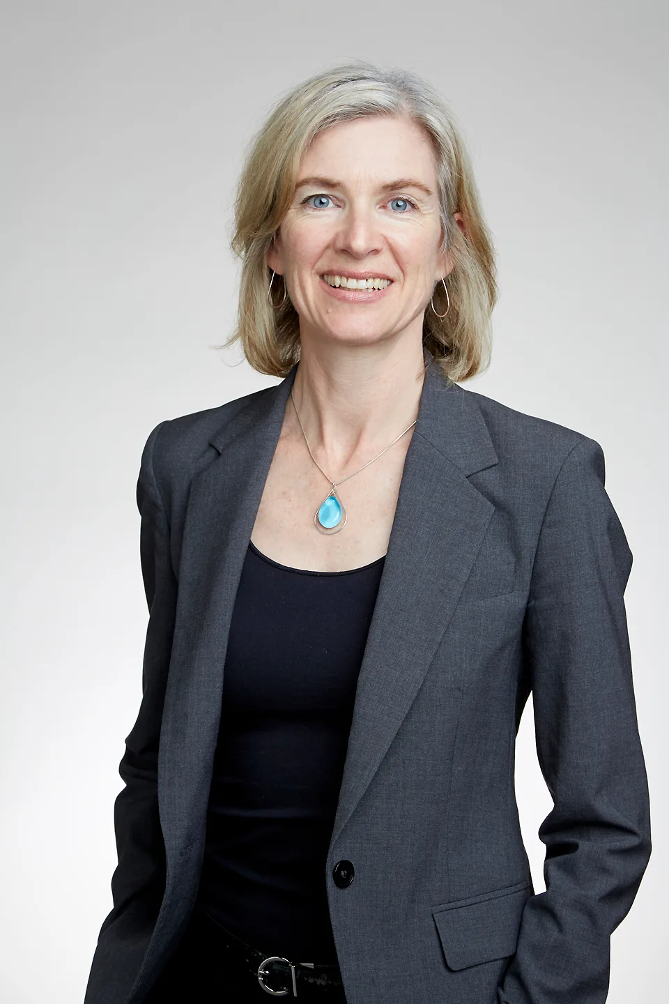 Jennifer Doudna'nın portresi: orta yaşlı bir kadın bilim insanı, omuzlarına dökülmüş düz sarı saçlar, koyu gömlek; nötr arka plan önünde sakin yüz ifadesi