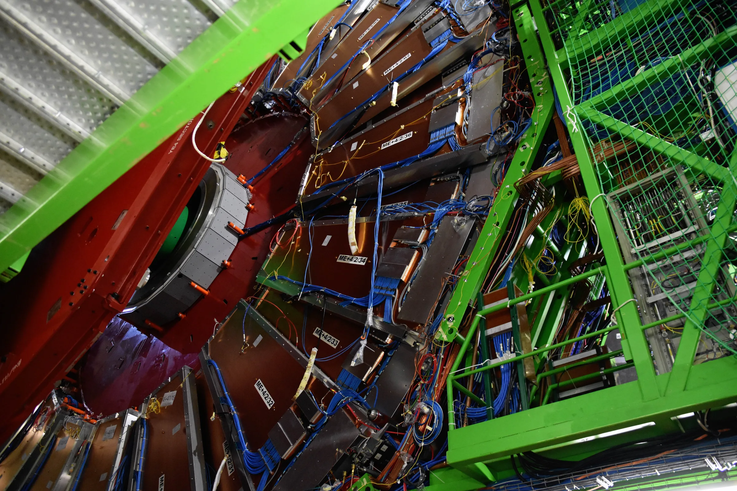 In a vast underground cavern, the Compact Muon Solenoid (CMS) detector seen from the open end: concentric steel rings, bundles of coloured cables and service platforms, with people-sized figures at the bottom for scale