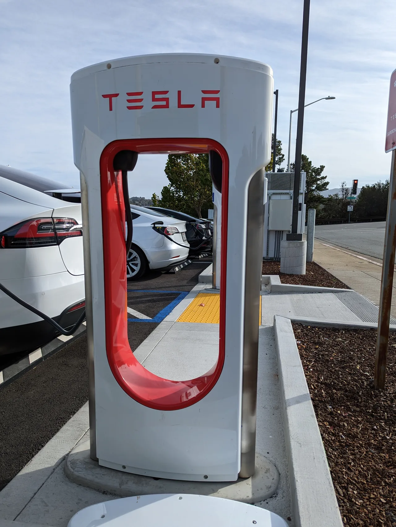 Kaliforniya San Mateo'da bir Tesla Süperşarjör istasyonunda şarj olan birden fazla Tesla otomobili — beyaz şarj direkleri ve gri zemin