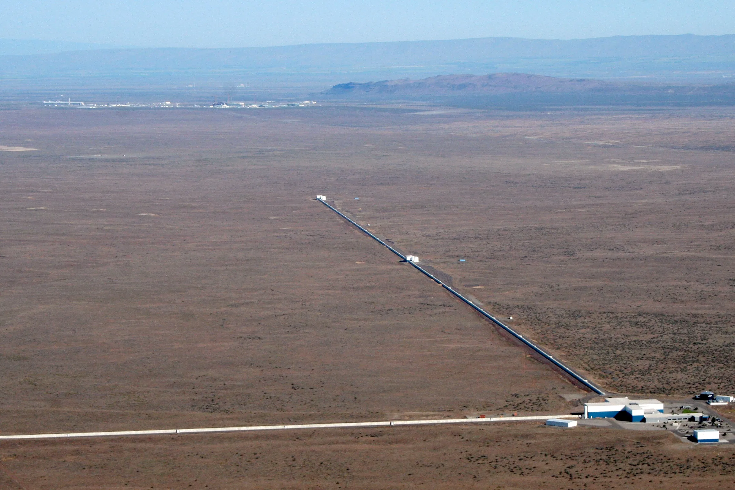 Aerial view of the LIGO Hanford observatory: two long concrete arms branching at right angles across desert terrain, each 4 km long