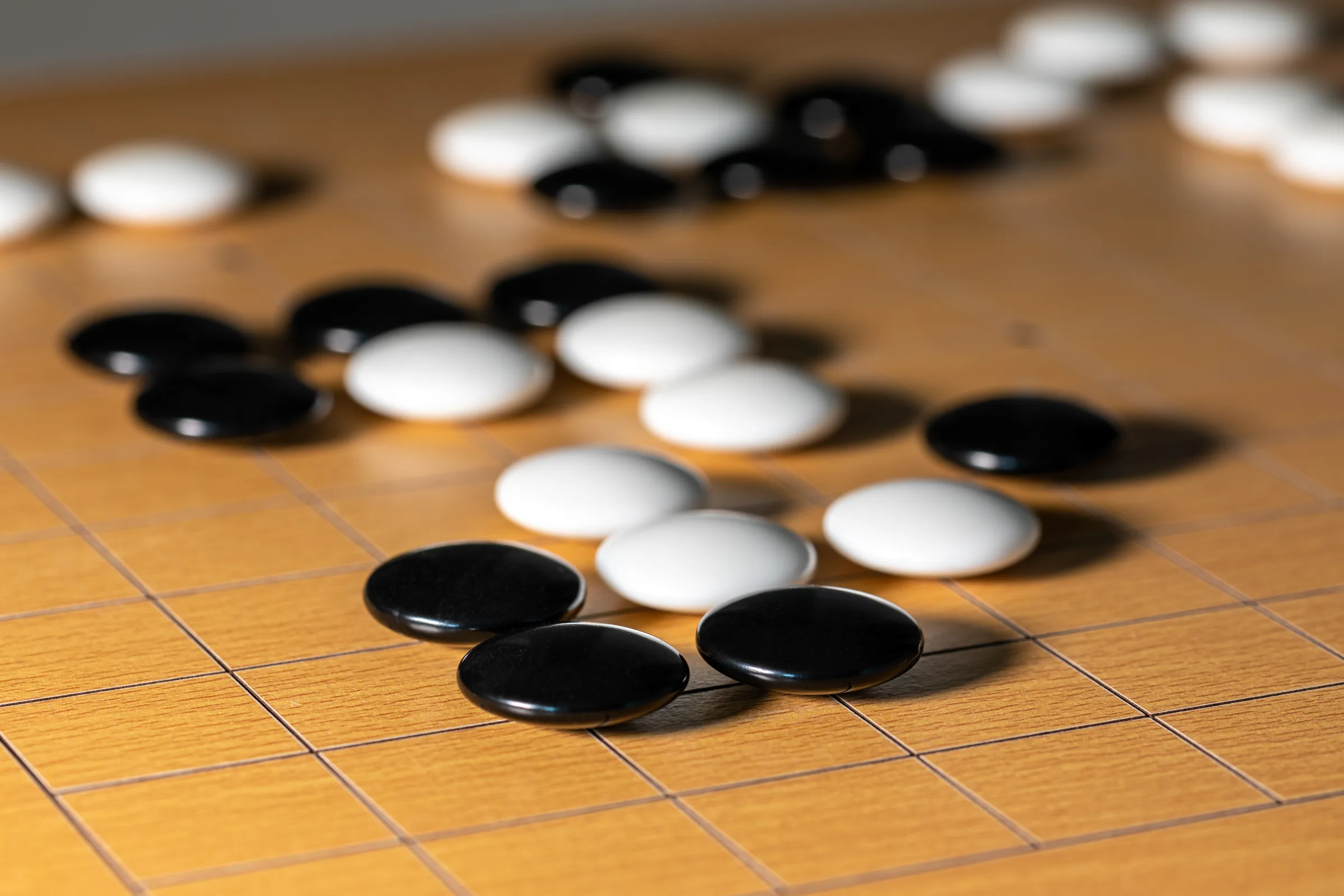 Close-up of a Go board: small white and black stones laid across the wooden surface, spare stones visible in a wooden bowl, light bringing out the grain of the board.