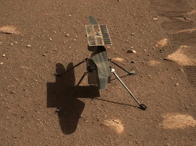 Ingenuity, a small four-legged helicopter, standing on the red soil and gravel of the Martian surface