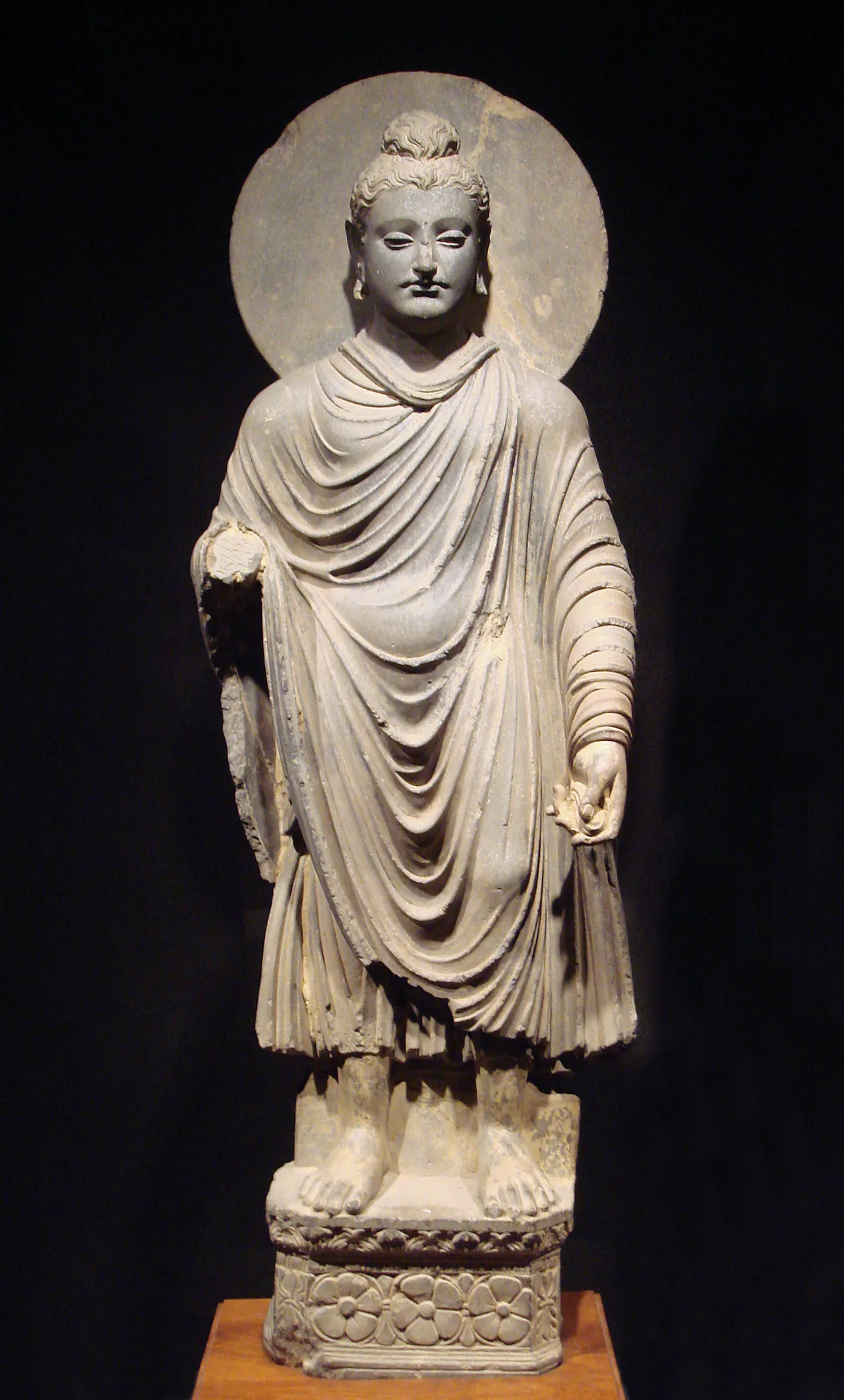 Standing gray schist Buddha statue showing Greco-Roman draped robes and wavy hair