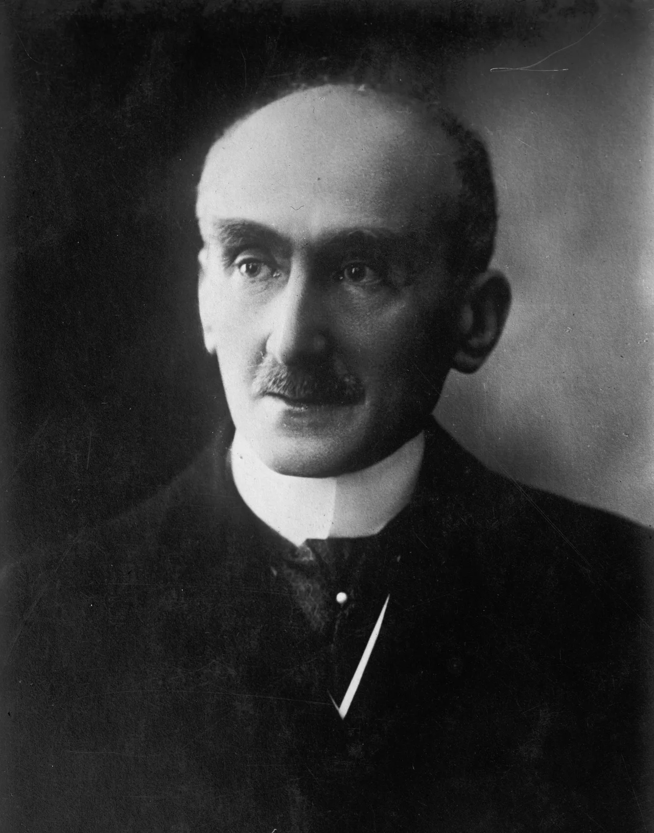 A c. 1927 photograph of Henri Bergson: a philosopher in a suit, leaning on a desk, with a white moustache, looking thoughtfully into the distance