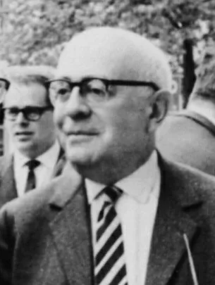 Photograph of Theodor Adorno: a serious, bespectacled man leaning at a desk against a wall