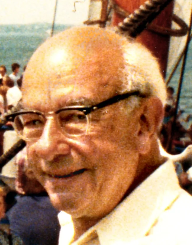 A 1980 color photograph of an older W. V. O. Quine, in a cap and glasses, on the deck of the schooner Bluenose II in Halifax harbour