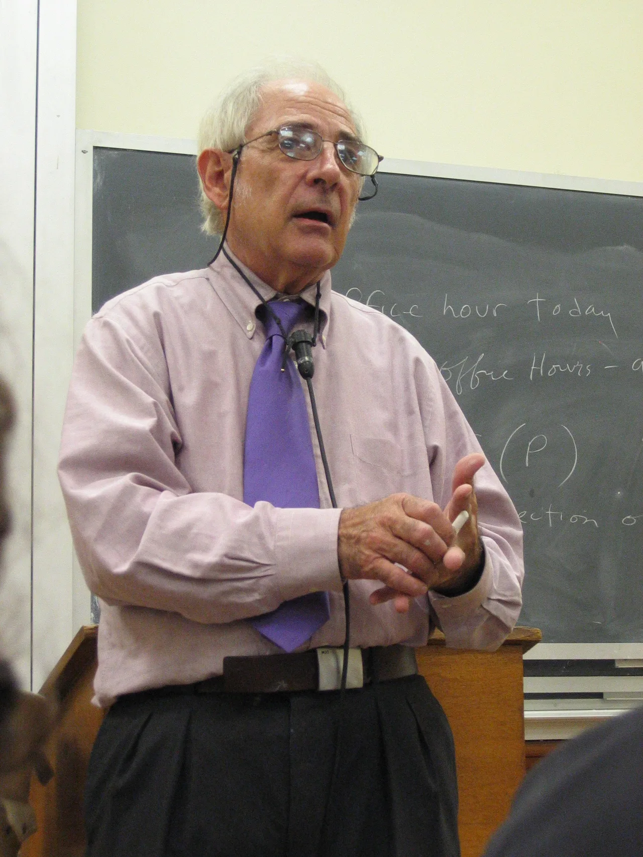 A photograph of John Searle: an older American philosopher with grey hair, speaking in an office setting