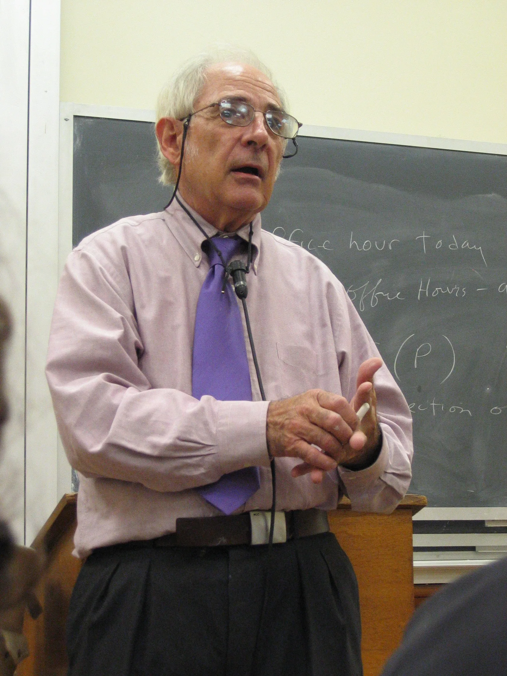 A photograph of John Searle: an older American philosopher with grey hair, speaking in an office setting