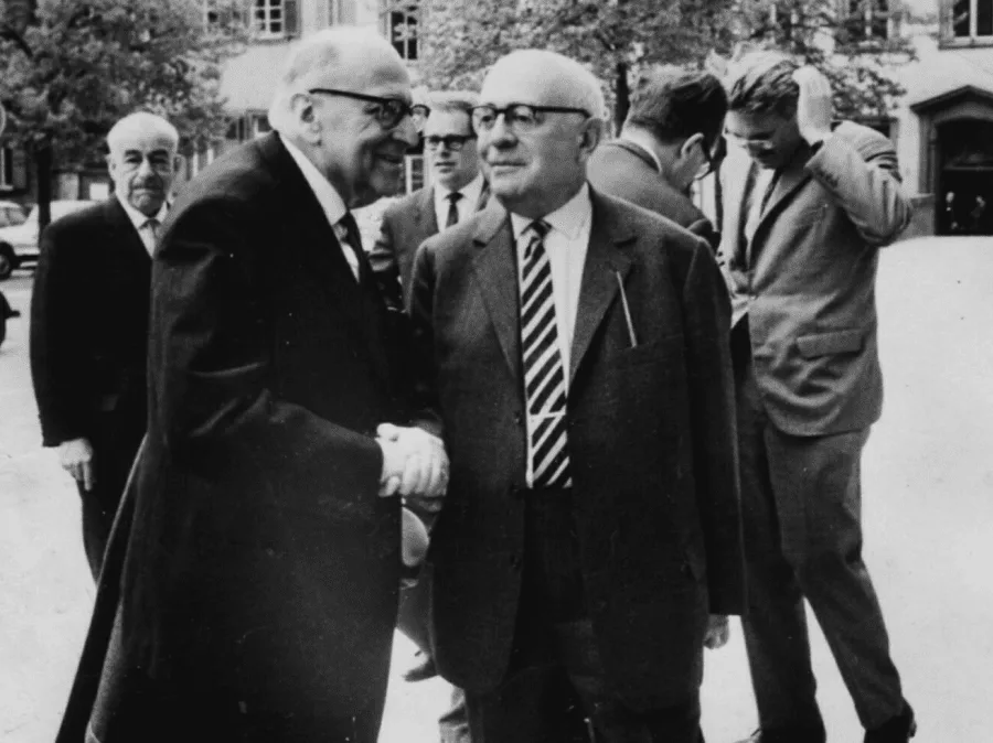 Heidelberg 1964: Theodor Adorno on the front right wearing glasses and holding a cigarette; Max Horkheimer leaning toward him on the front left; the young Jürgen Habermas in the background right, running his hand through his hair
